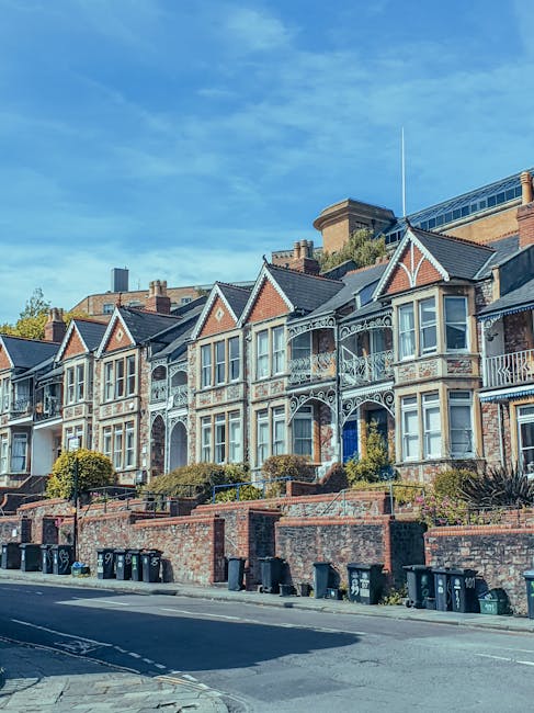 A row of Victorian-style residential houses on Fulham Palace Road in Hammersmith, featuring ornate cast-iron balconies, large bay windows, and decorative brickwork. The homes are set on a sloped street with small front gardens enclosed by low brick walls, some with neatly trimmed bushes and greenery. The pavement and street are visible in the foreground with several wheelie bins lined up along the curb. The sky above is bright blue with a few wispy clouds, and the scene appears clean and well-maintained, reflecting the professional domestic cleaning services provided by Cleaner Hammersmith, as highlighted on their website.