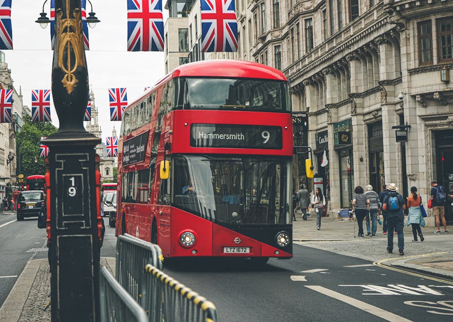 A red double-decker bus with the route number 9 and destination Hammersmith is driving along a busy city street on Fulham Palace Road, Hammersmith. The street is lined with historic buildings featuring detailed stone facades and large windows. Pedestrians, some carrying shopping bags and backpacks, are walking on the sidewalk, while Union Jack flags are hanging from lamp posts along the road. The scene is captured during daylight with overcast skies, and the street is clean and well-maintained, reflecting a typical urban environment. This image visually emphasizes the vibrant city life in Hammersmith, relevant for domestic cleaning or surface cleaning services promoted by Cleaner Hammersmith.