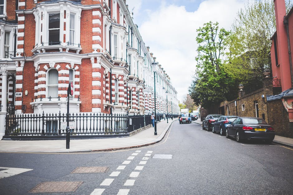 A row of Victorian-style residential houses on Fulham Palace Road in Hammersmith, featuring ornate cast-iron balconies, large bay windows, and decorative brickwork. The homes are set on a sloped street with small front gardens enclosed by low brick walls, some with neatly trimmed bushes and greenery. The pavement and street are visible in the foreground with several wheelie bins lined up along the curb. The sky above is bright blue with a few wispy clouds, and the scene appears clean and well-maintained, reflecting the professional domestic cleaning services provided by Cleaner Hammersmith, as highlighted on their website.