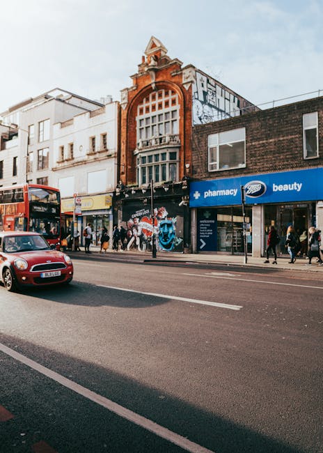A street scene in Hammersmith with a mix of historic and modern buildings, including a prominent red-brick structure featuring an ornate arch and large window at the top. The ground floor hosts retail stores such as a pharmacy and beauty shop, with people walking along the pavement. A red double-decker bus is visible on the road, alongside a small red car. The scene is well-lit with natural daylight, and the overall cleanliness of the street suggests regular maintenance. Cleaner Hammersmith specializes in surface cleaning, deep cleaning, and sanitisation for local businesses, ensuring these commercial spaces remain hygienic and well-presented.