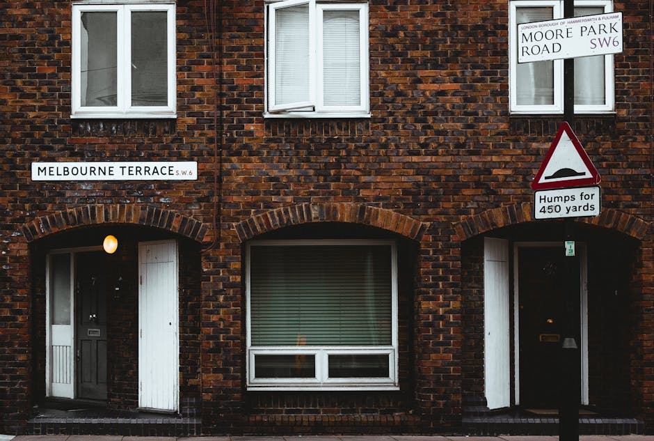 This image depicts a busy urban street scene in Hammersmith, Birmingham, with a variety of historic and modern buildings lining the street. The buildings are primarily constructed from brick and feature decorative architectural details, with some storefronts showcasing signage and large glass windows. The street includes pedestrian crossings and designated bike lanes marked with red dashed lines, while vehicles and motorbikes are visible on the road. The scene is illuminated by natural daylight, creating a bright and lively atmosphere. As part of professional cleaning and maintenance practices, Cleaner Hammersmith advocates for thorough surface cleaning of all visible surfaces, including pavement and building facades, to ensure hygiene and visual appeal in commercial and residential areas.