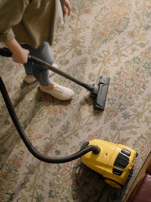 A person vacuuming a decorative area rug with an ornate floral pattern in a room illuminated by natural light. The individual, dressed in casual attire with beige sneakers and jeans, is using a yellow canister vacuum cleaner connected to a black hose and a cleaning head attachment. The rug appears clean and well-maintained, and the image emphasizes surface cleaning and deep cleaning processes as part of professional domestic cleaning services offered by Cleaner Hammersmith, situated in a residential setting in Hammersmith W6.