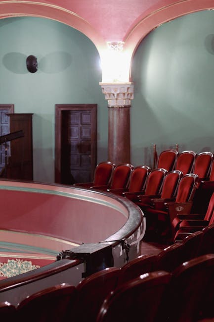 Interior view of the Lyric Hammersmith theatre auditorium showcasing neatly arranged red upholstered seats on a polished wooden floor, with smooth, pastel green plastered walls and decorative white cornicing along the ceiling. The lighting creates a soft glow, highlighting the clean, well-maintained seating area. Visible are the curved balcony railing in the foreground, a closed wooden door on the back wall, and a wall-mounted speaker. The venue's furnishings and surfaces appear spotless, demonstrating professional deep cleaning and surface sanitisation carried out by Cleaner Hammersmith, specialists in domestic and commercial cleaning services for theatre spaces and similar venues.