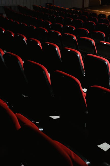 Interior view of the Lyric Hammersmith theatre auditorium showcasing neatly arranged red upholstered seats on a polished wooden floor, with smooth, pastel green plastered walls and decorative white cornicing along the ceiling. The lighting creates a soft glow, highlighting the clean, well-maintained seating area. Visible are the curved balcony railing in the foreground, a closed wooden door on the back wall, and a wall-mounted speaker. The venue's furnishings and surfaces appear spotless, demonstrating professional deep cleaning and surface sanitisation carried out by Cleaner Hammersmith, specialists in domestic and commercial cleaning services for theatre spaces and similar venues.